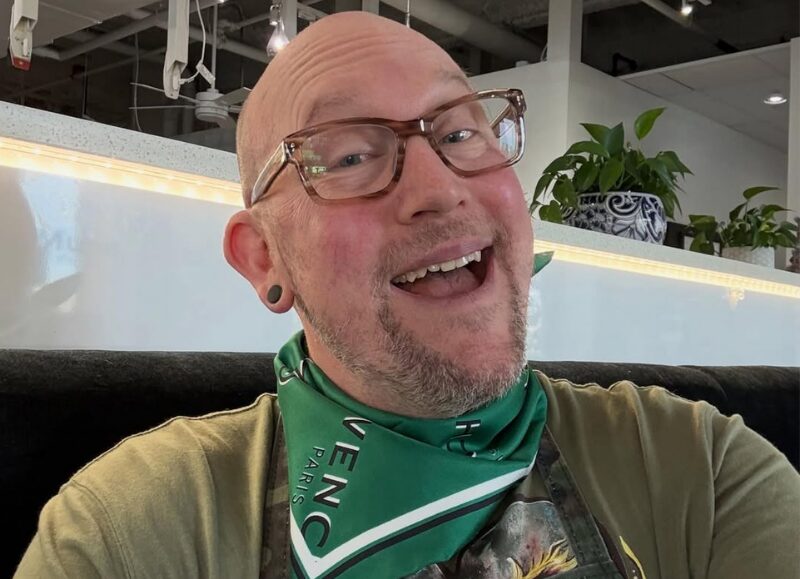 Close-up of smiling educator Todd Faulk wearing glasses and a green Givenchy neck scarf inside the Aveda Arts Dallas campus.
