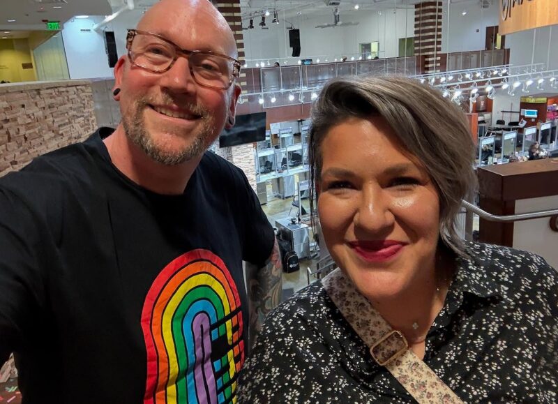 Selfie of Todd Faulk wearing a rainbow graphic shirt standing alongside a smiling colleague overlooking the salon floor at Aveda Arts Dallas.