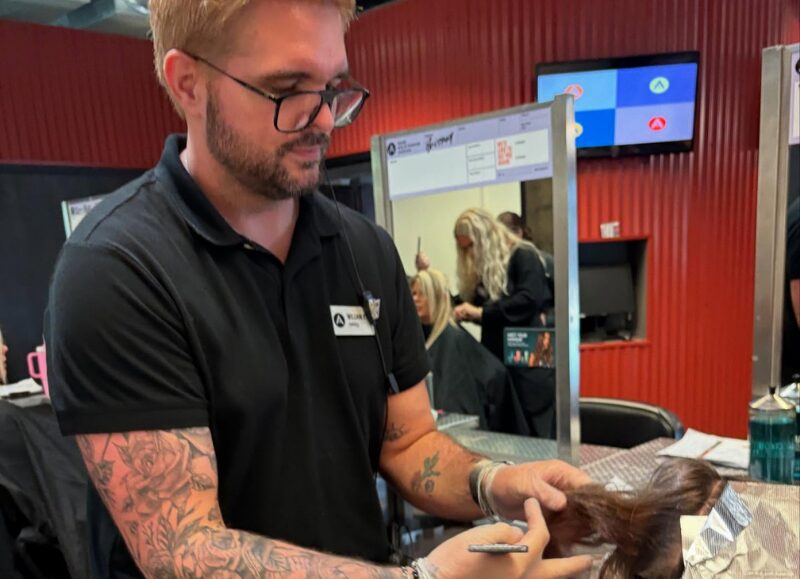 Aveda Arts Educator William Richard, showing off his arm tattoos, wears black nitrile gloves while carefully sectioning a mannequin's brown hair with foil during a foiling technique demonstration on a salon campus.