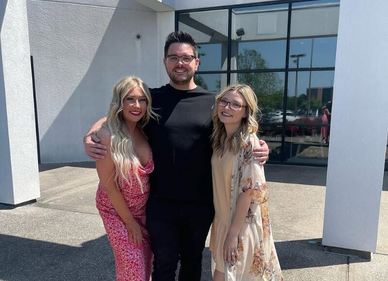 Three people smiling widely together on a sunny outdoor patio, with a man in the center arm-in-arm with two women. The photo captures the diverse and supportive atmosphere that defines the Aveda Arts cosmetology school culture.