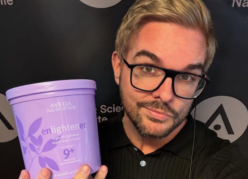 Selfie portrait of Aveda Arts Educator William Richard with blonde hair and black glasses, holding a large purple tub of "AVEDA enlighten super éclaircissant high-lift lightener 9+" in front of an Aveda-branded step-and-repeat banner.