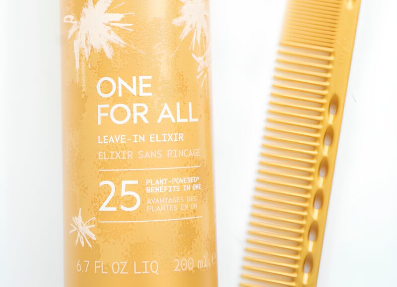 Aveda One For All Elixir alongside a golden cutting and detangling comb on a clean white surface.