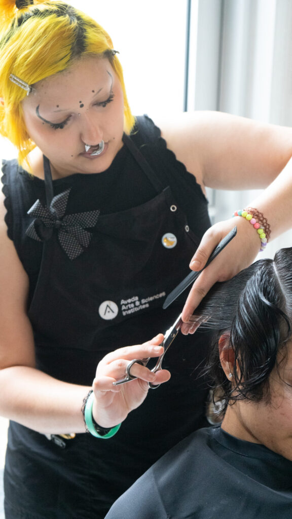 Aveda Arts student with bright yellow hair using shears and a comb to cut a section of a guest’s wet hair.