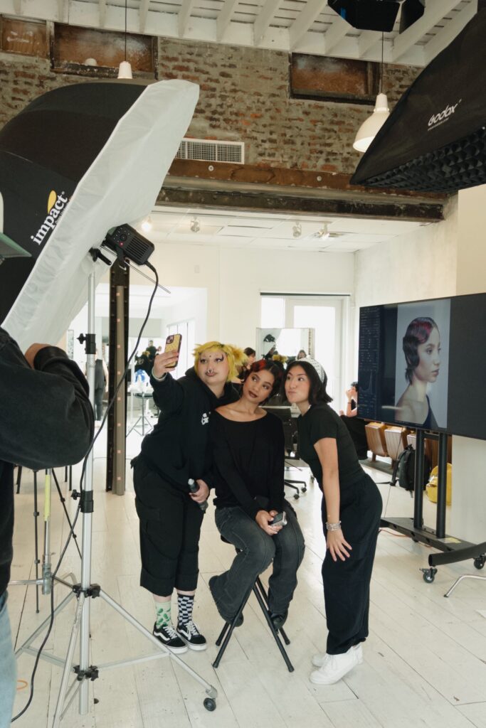 Three people taking a selfie beside studio lights and a monitor displaying a finished hairstyle.